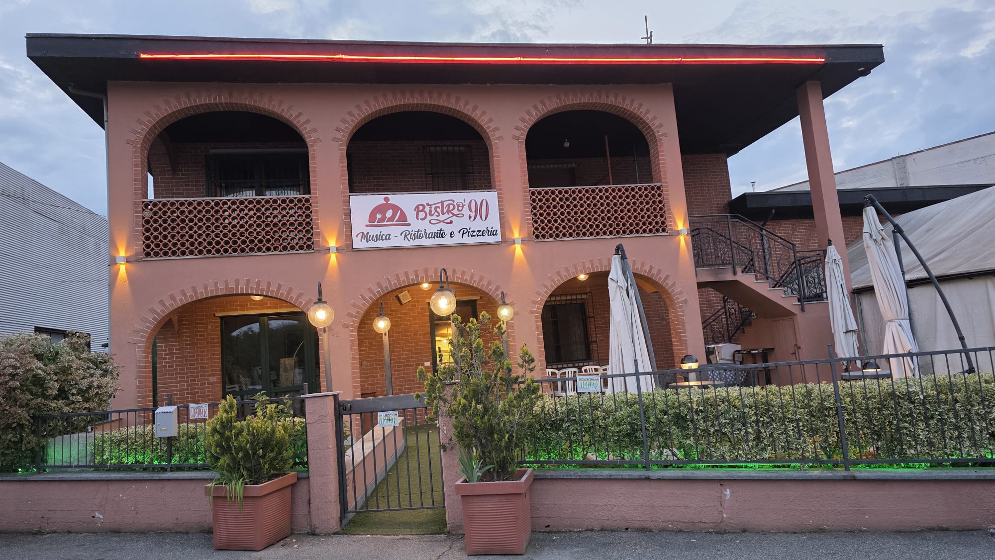 Facciata rosa ad archi con insegna «Bistro' 90 Musica - Ristorante e Pizzeria», neon rosso, siepe e ombrelloni chiusi