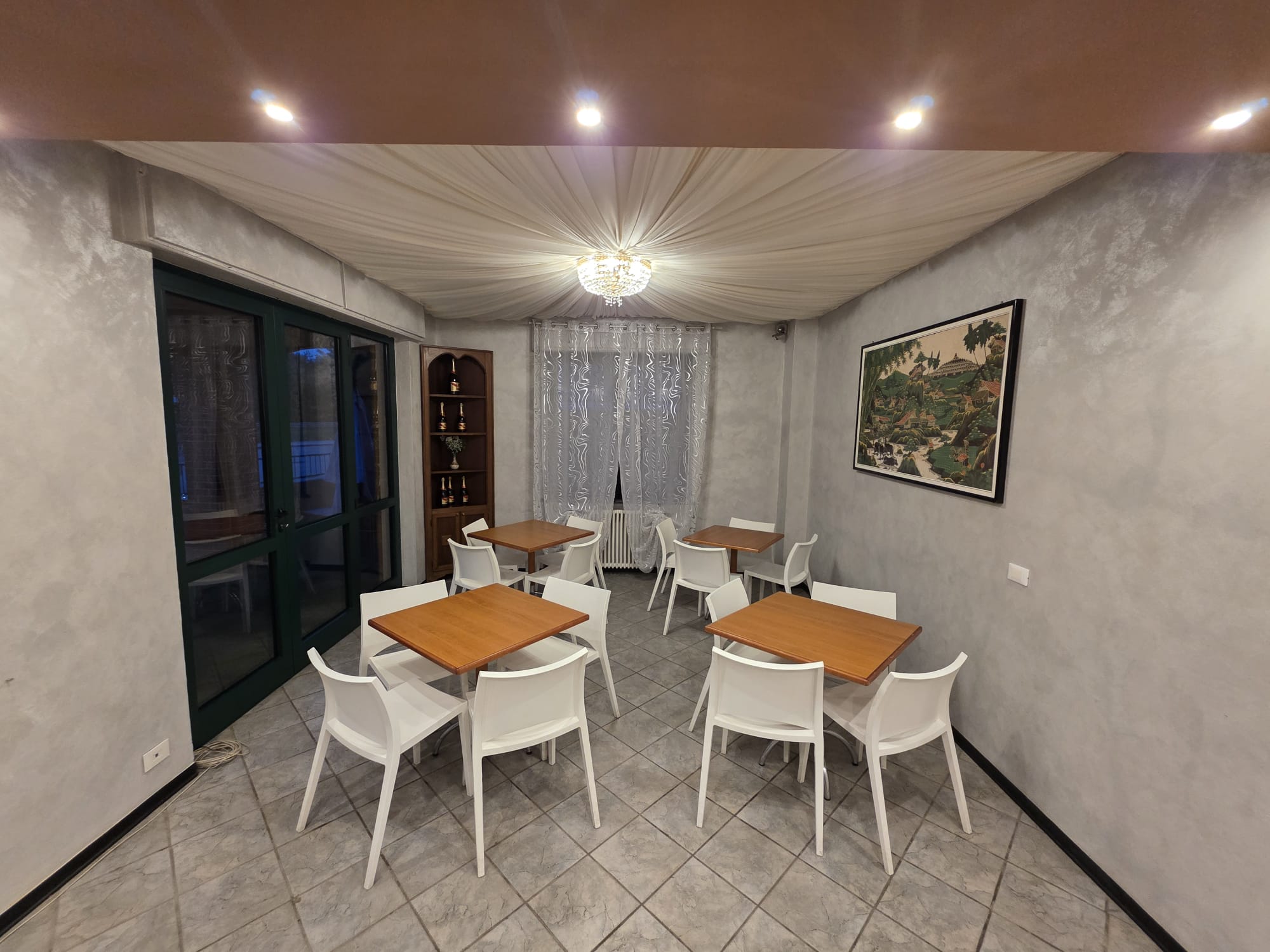 Sala di Bistro' 90 con tavoli in legno, sedie bianche, soffitto drappeggiato, lampadario di cristallo e quadro alla parete